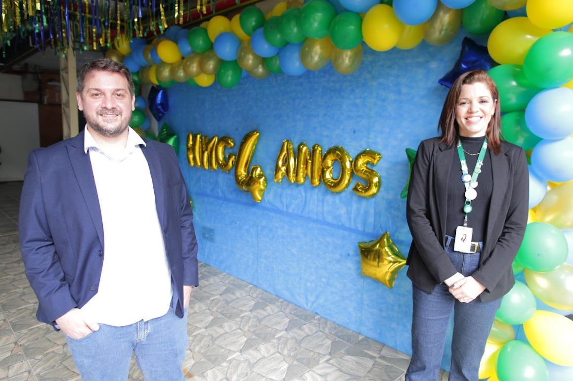 A foto mostra um homem e uma mulher lado a lado, de pé; eles estão sorrindo e posando em frente a uma parede azul decorada com balões azuis, verdes e amarelos e na qual se lê “HMG 4 anos” em letras douradas